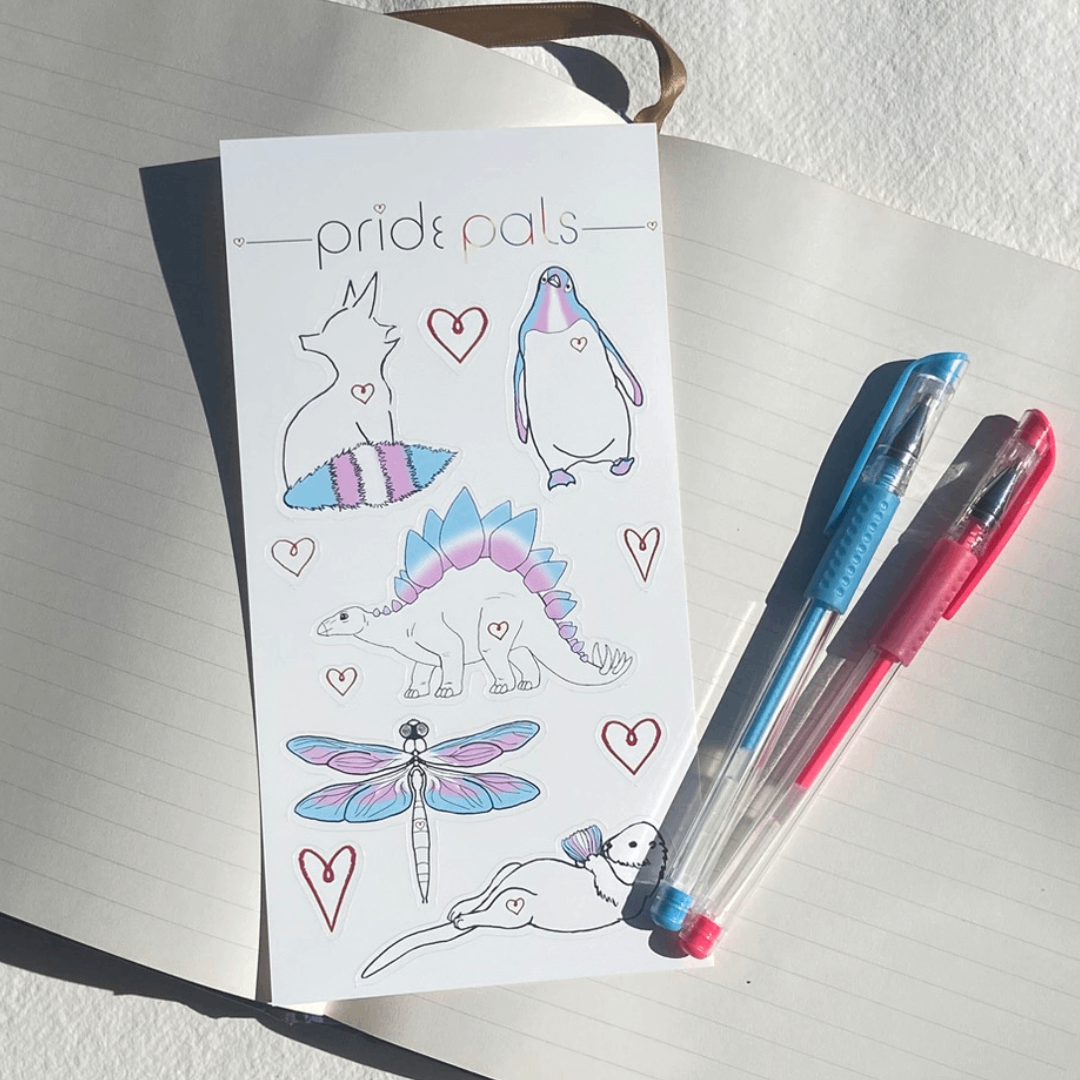 Sheet of vinyl stickers with animal designs and 'pride pals' branding, placed on an open notebook with two pens.